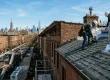Two roofers installing asphalt shingles on a Brooklyn brownstone with the Manhattan skyline in the background.