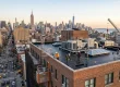 roofing crew working on a flat brick building roof in Manhattan, with a rooftop crane hoisting materials and a dense NYC cityscape and traffic-filled street in the background.